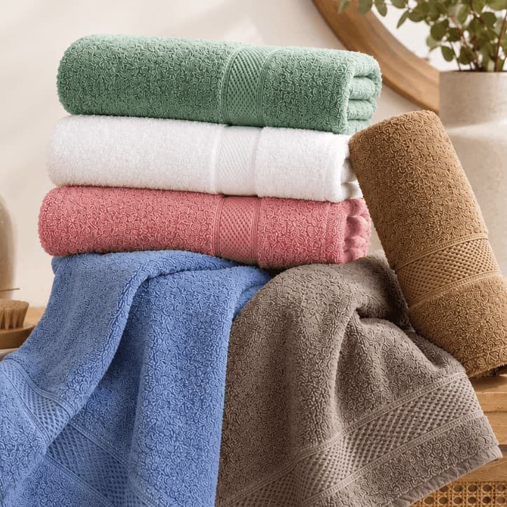 Sáu chiếc khăn tắm Toweland màu xanh lá, trắng, hồng, xanh dương, ghi và nâu, xếp chồng và vắt, phía sau có gương tròn và chậu cây.